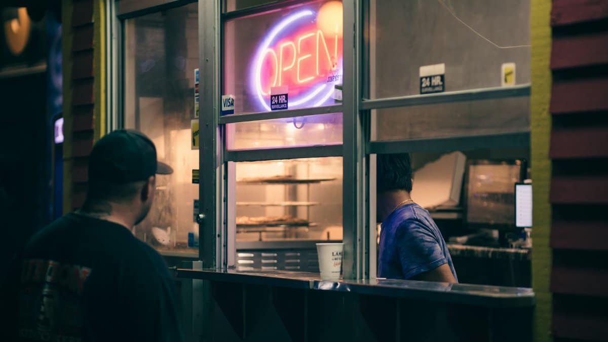 Man ordering food at a street stall with neon 'Open' sign at night.