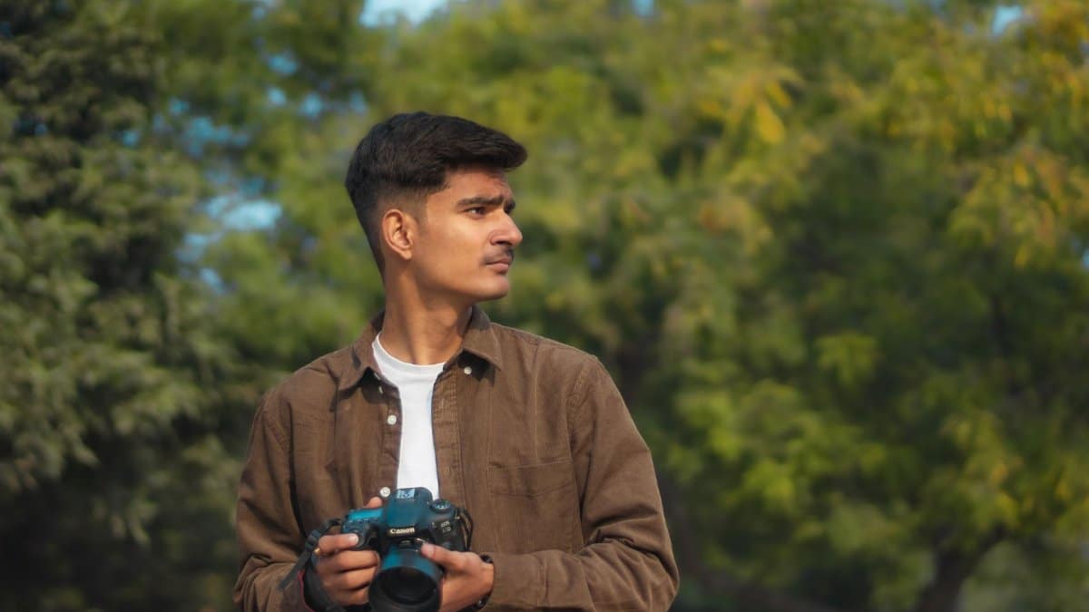 Young male photographer holding a camera in a green outdoor setting, looking thoughtful.