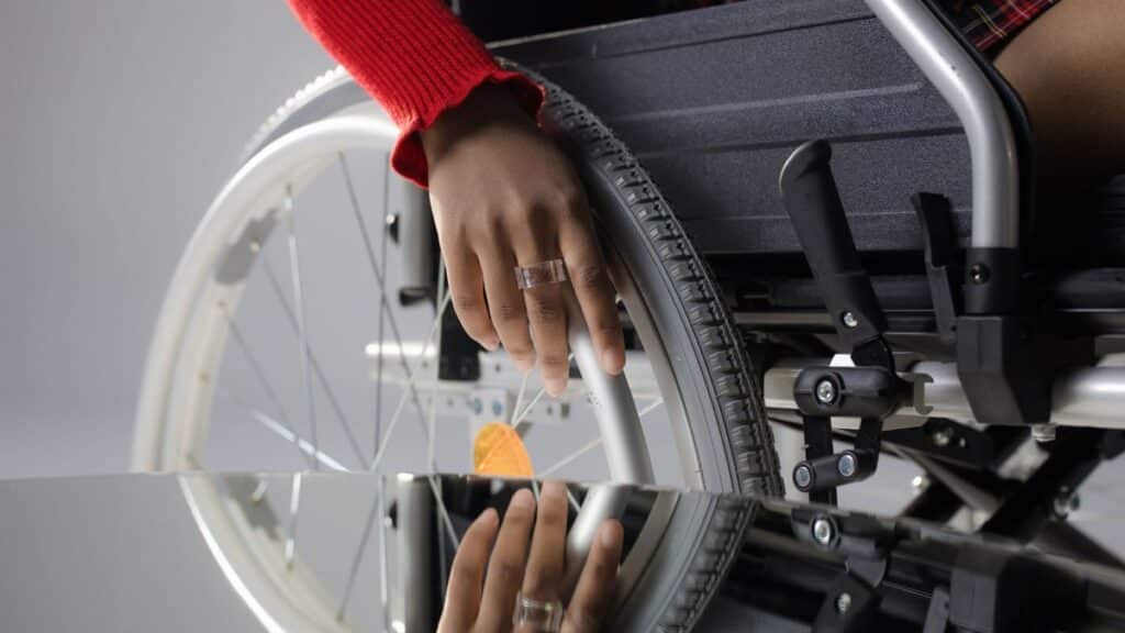 Close-up of a person's hand on a wheelchair wheel, reflected in glass, highlighting mobility and disability themes.