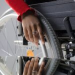 Close-up of a person's hand on a wheelchair wheel, reflected in glass, highlighting mobility and disability themes.