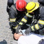 Firefighters provide CPR to an injured man using a bag valve mask on a cobblestone street.