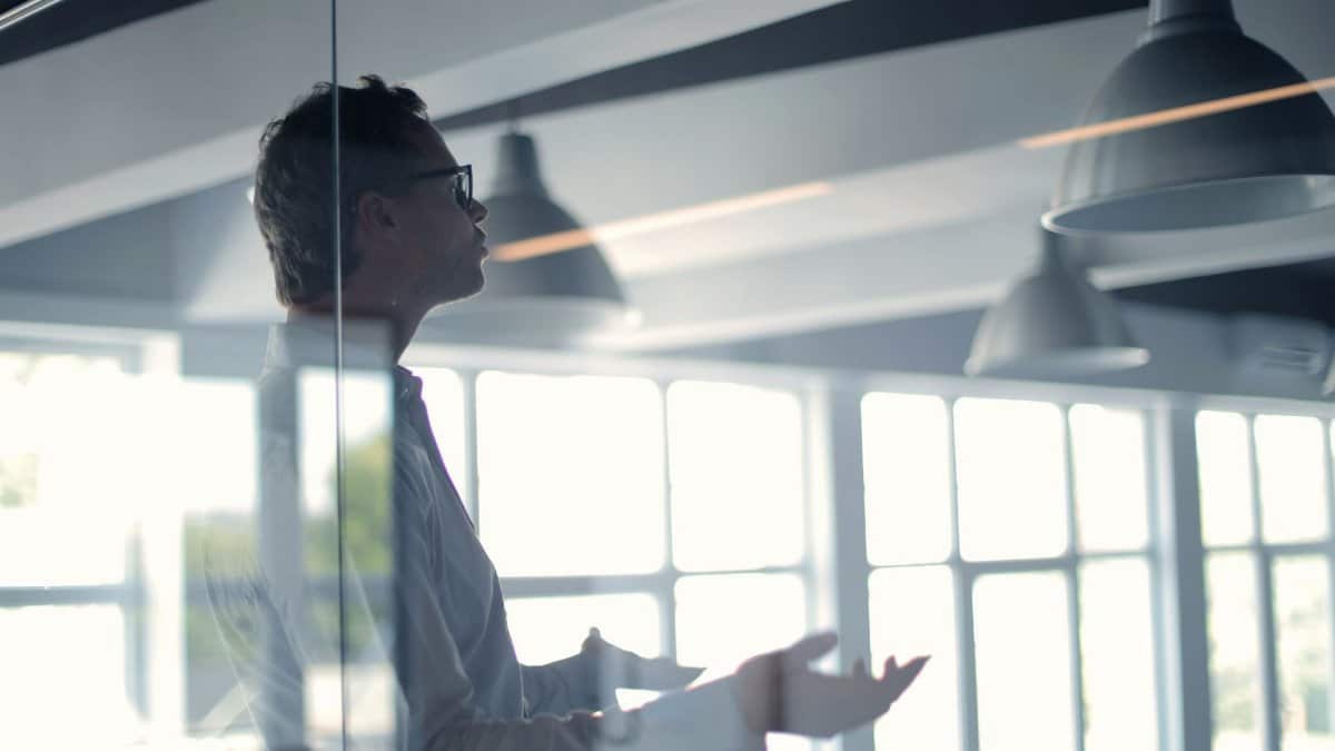 Through glass view of adult businessman in glasses and white shirt gesturing while holding presentation in spacious office