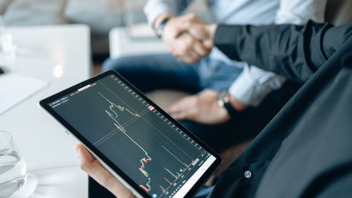 Two adults shaking hands with a stock market chart on a tablet, symbolizing a business deal.
