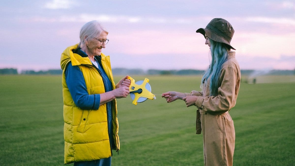 Elderly and young woman using a measuring tool in a grassy field during sunset.
