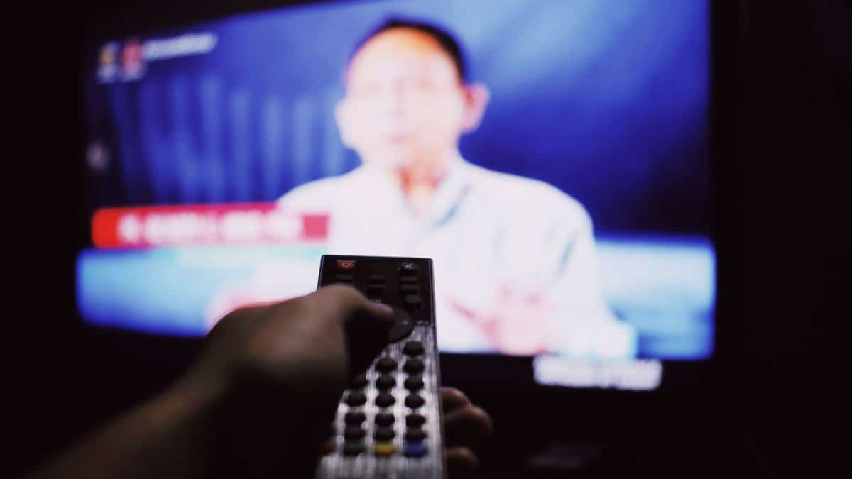 Crop anonymous person with remote controller switching channels while watching TV on blurred background in living room with dim light
