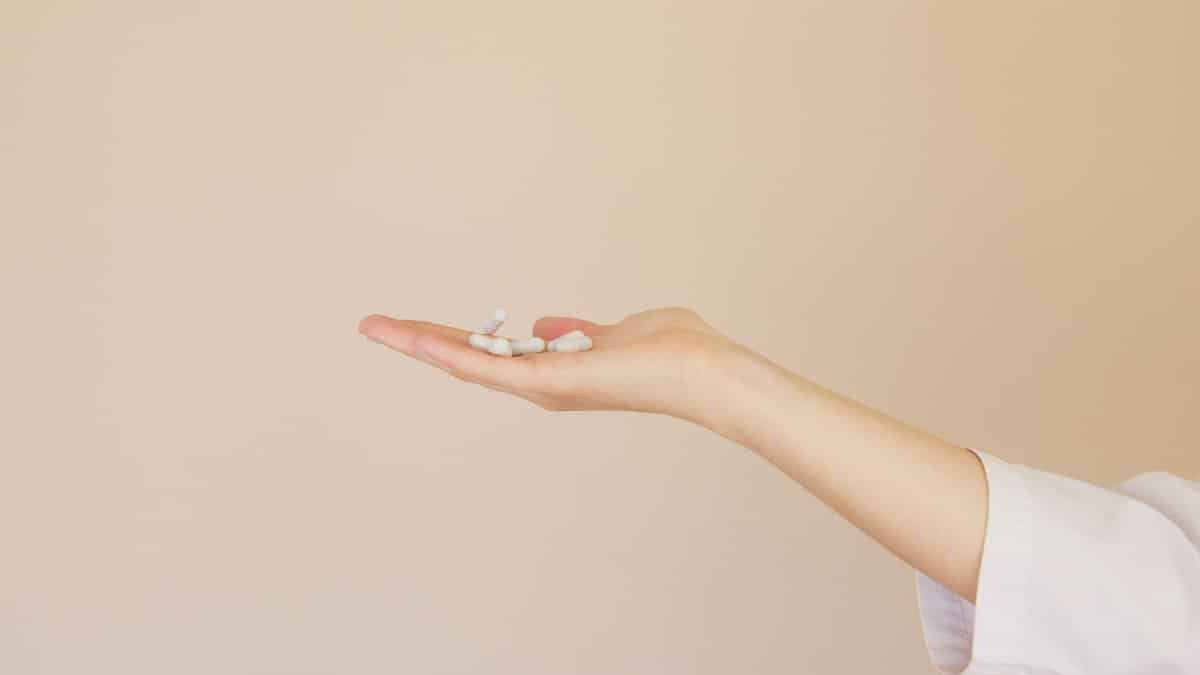 Side view of crop unrecognizable female pharmaceutist in white uniform with pile of white painkillers on palm against light brown background