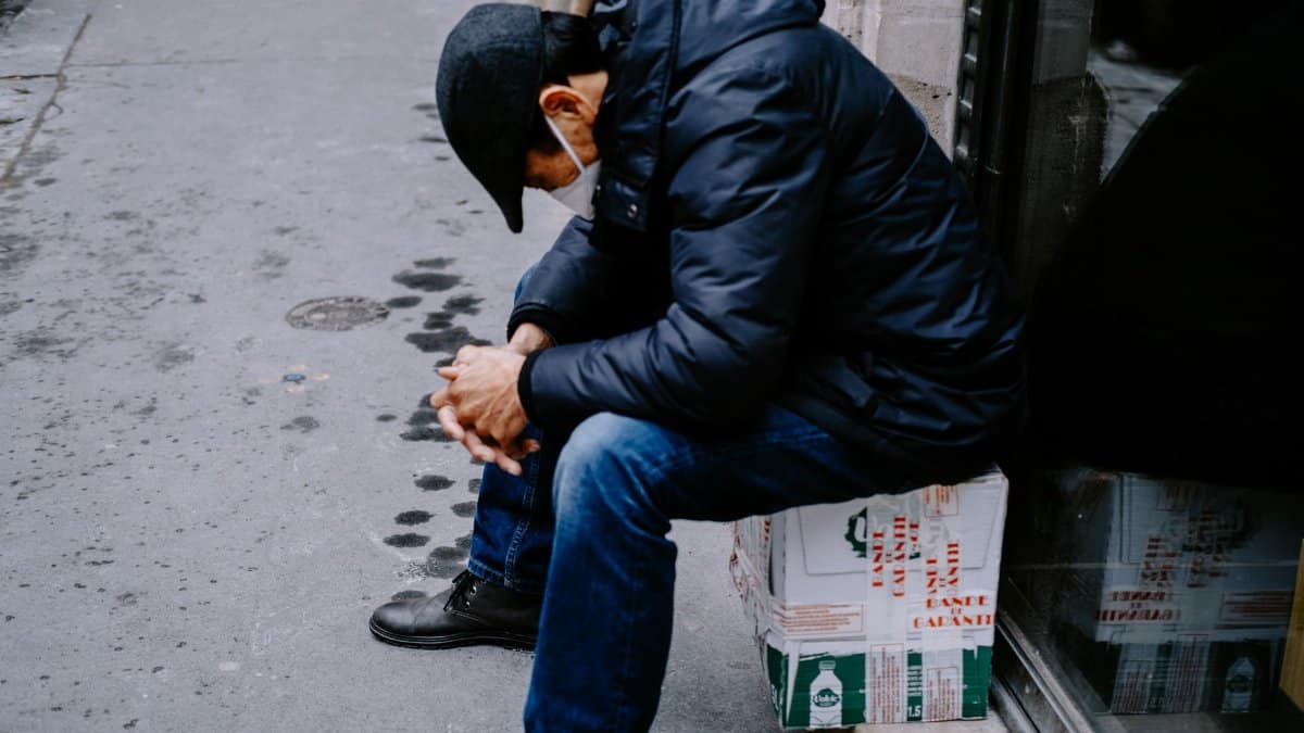A man wearing a face mask sits on a box in an urban setting, looking pensive.