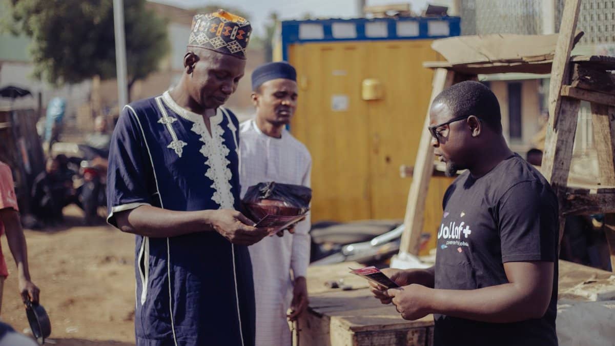 Three men engage in conversation at an outdoor market with traditional and modern attire.