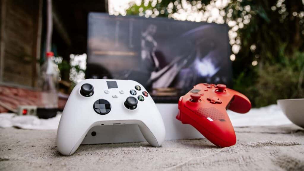 White and red Xbox wireless controllers set outdoors. A computer screen is blurred in the background.