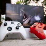 White and red Xbox wireless controllers set outdoors. A computer screen is blurred in the background.