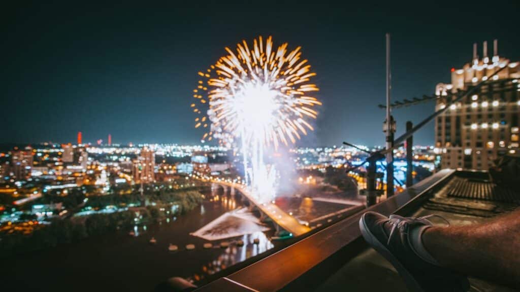 Fireworks illuminate the night sky over Minneapolis cityscape, viewed from a rooftop.