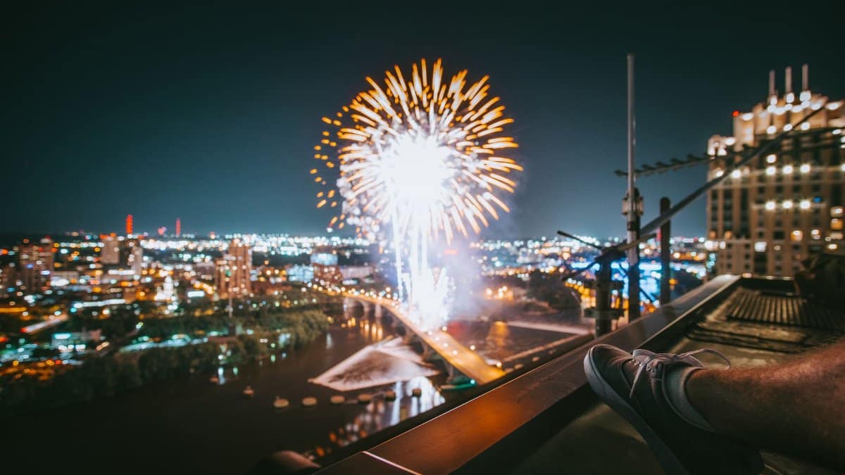 Fireworks illuminate the night sky over Minneapolis cityscape, viewed from a rooftop.