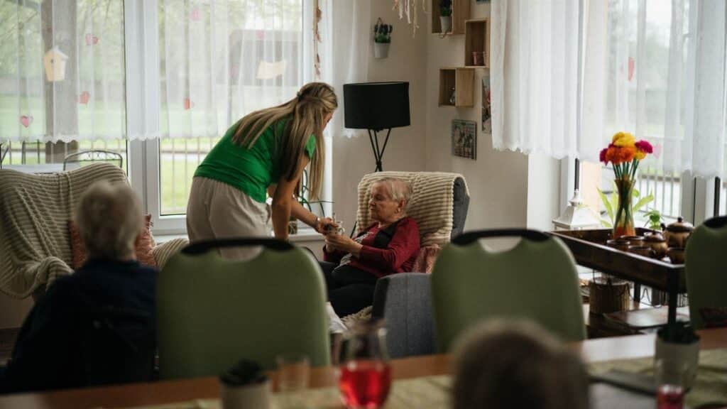 Caregiver assisting elderly women with tea in a cozy nursing home setting.
