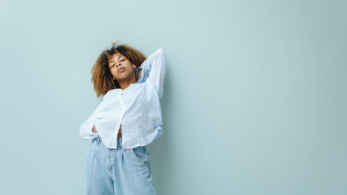 Fashionable woman standing confidently with afro hair and denim, against a light blue background.