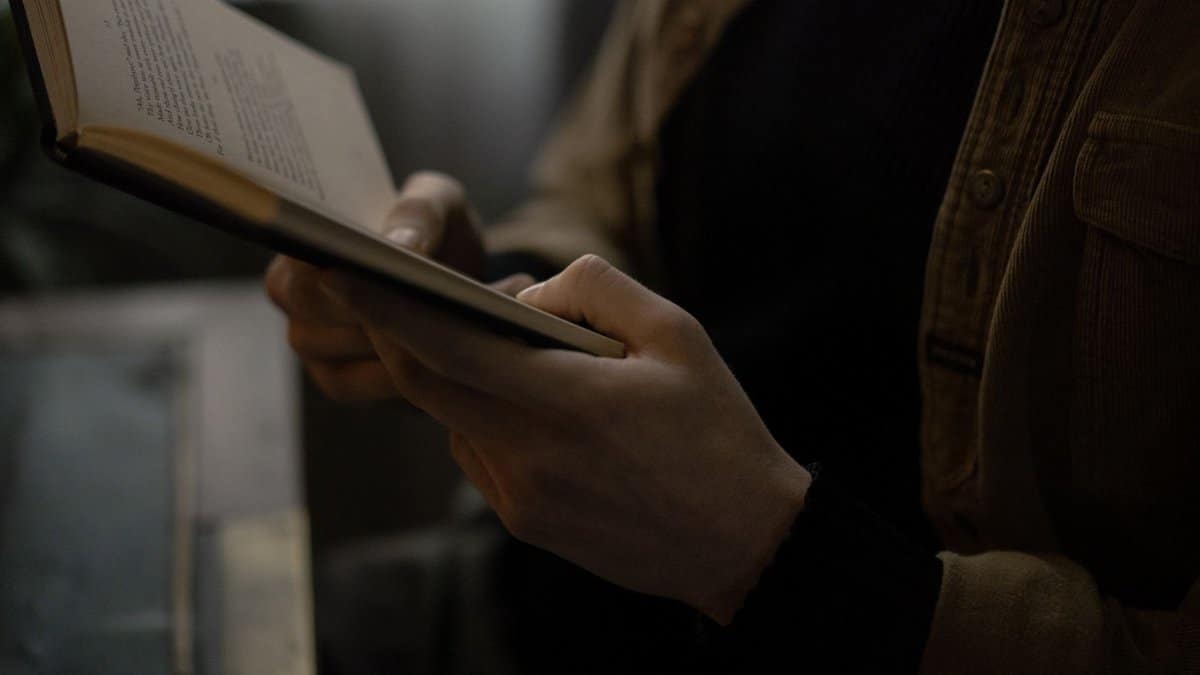 A person in a brown jacket holds an open book, creating a cozy reading atmosphere indoors.
