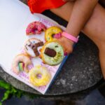 A person holds a decorated doughnut from a colorful assortment in a box outdoors.