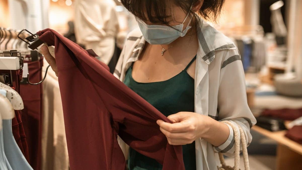 A woman shops indoors wearing a face mask, examining clothes during the pandemic era.