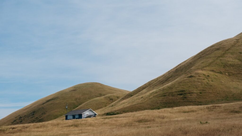 Tranquil countryside landscape with a small house nestled between rolling hills under a blue sky.