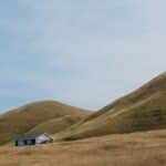 Tranquil countryside landscape with a small house nestled between rolling hills under a blue sky.