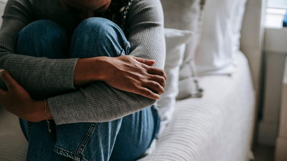Crop unhappy mournful African American female wearing casual outfit embracing knees on bed and hiding face in sad thoughts