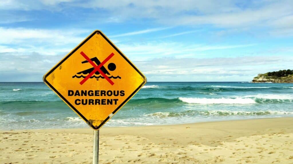 Yellow warning sign indicating dangerous currents at Bondi Beach, Australia, with ocean waves in the background.