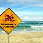Yellow warning sign indicating dangerous currents at Bondi Beach, Australia, with ocean waves in the background.