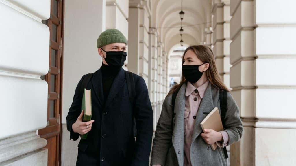 Two students in face masks holding books walking through a historic hallway, showcasing new normal lifestyle.
