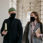 Two students in face masks holding books walking through a historic hallway, showcasing new normal lifestyle.