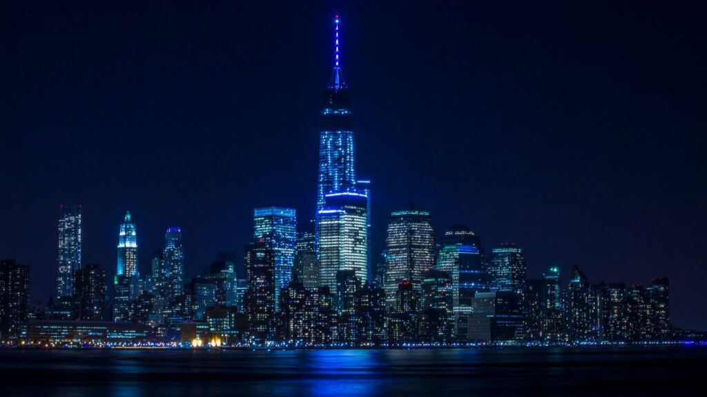 Illuminated New York City skyline reflecting over the Hudson River at night.
