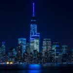 Illuminated New York City skyline reflecting over the Hudson River at night.
