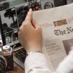 Close-up of a person reading The New York Times by a laptop and camera.