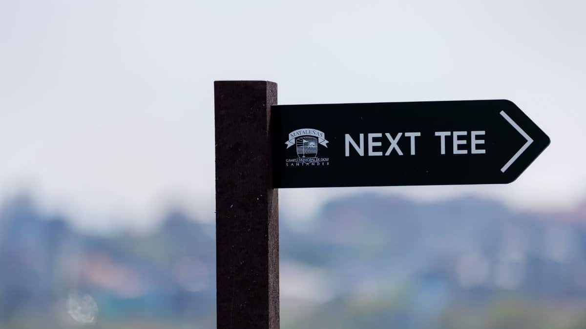 A directional sign on a golf course pointing to the next tee against a blurred background.
