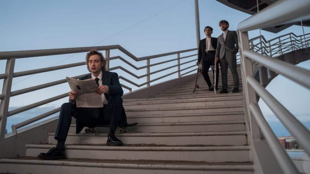 Three stylish men on a staircase, one reading a newspaper, others holding skateboards.