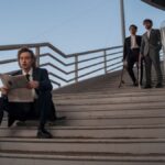 Three stylish men on a staircase, one reading a newspaper, others holding skateboards.