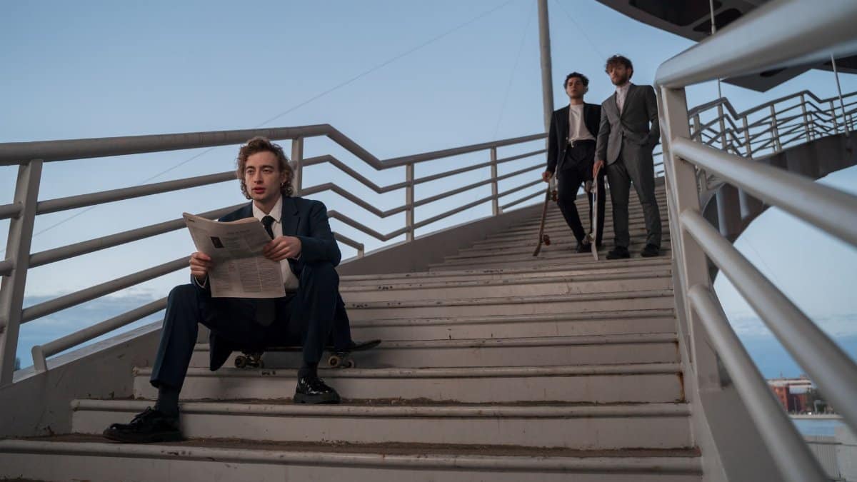 Three stylish men on a staircase, one reading a newspaper, others holding skateboards.