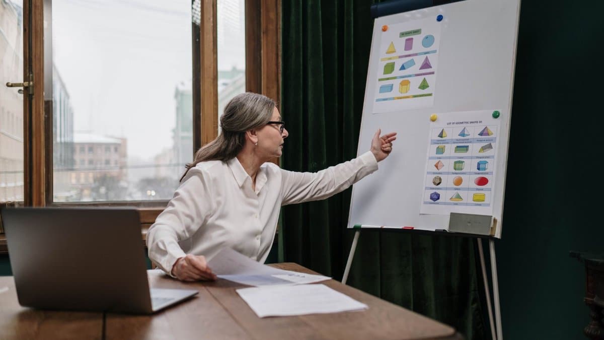 Senior woman conducting an online geometry lesson, highlighting various shapes on a chart with a laptop.