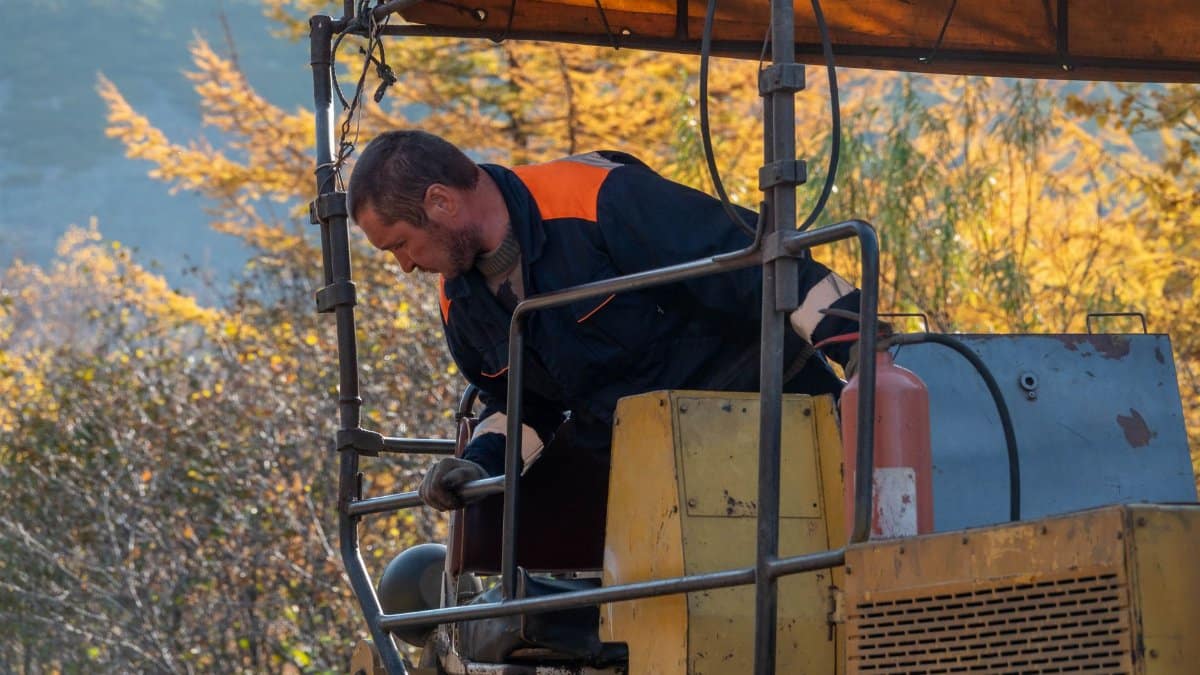 A construction worker in a blue uniform operating heavy machinery outdoors during autumn.