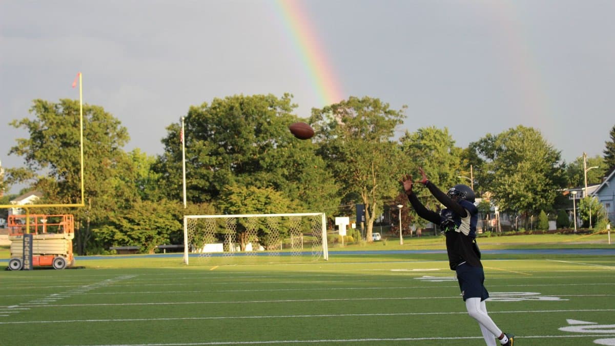Football player leaps to catch a ball with a vibrant rainbow backdrop on a sunny field.