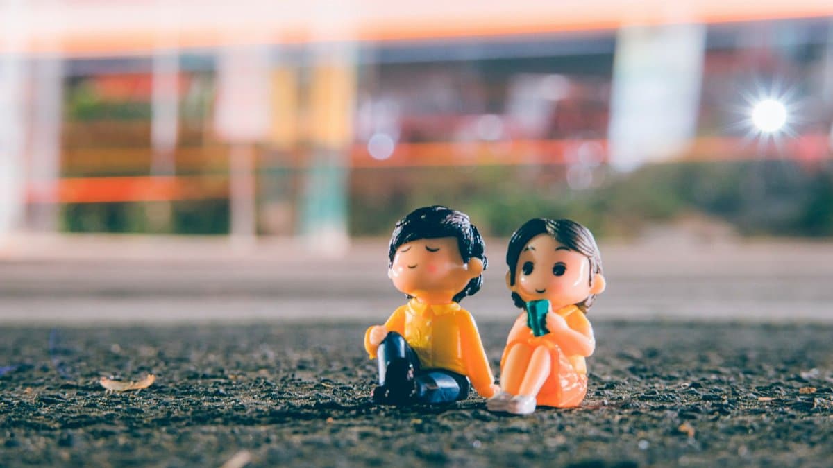 Ground level of small figurines of boy and girl placed on asphalt walkway near roadway with long exposure light in evening time on blurred background
