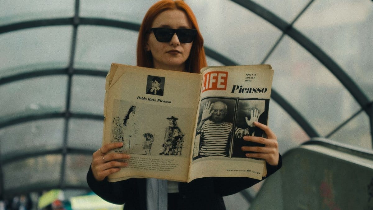 A woman with red hair and sunglasses reads a Life magazine about Picasso in an urban subway setting.