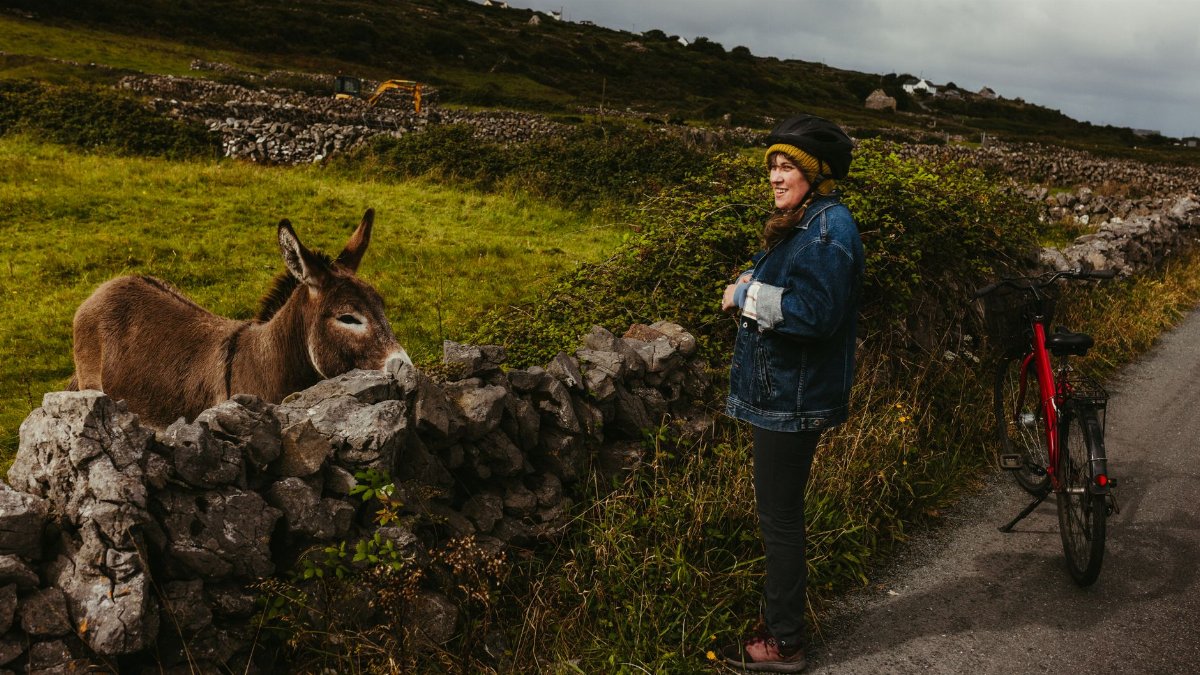 A woman interacts with a donkey near a stone wall on Inishmore, Galway, Ireland, with a bicycle beside her.