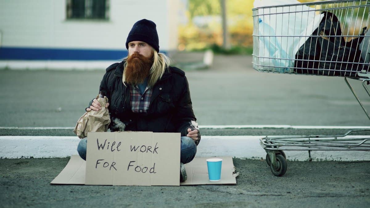 Man sitting outdoors with a sign reading 'Will work for food', next to shopping cart.