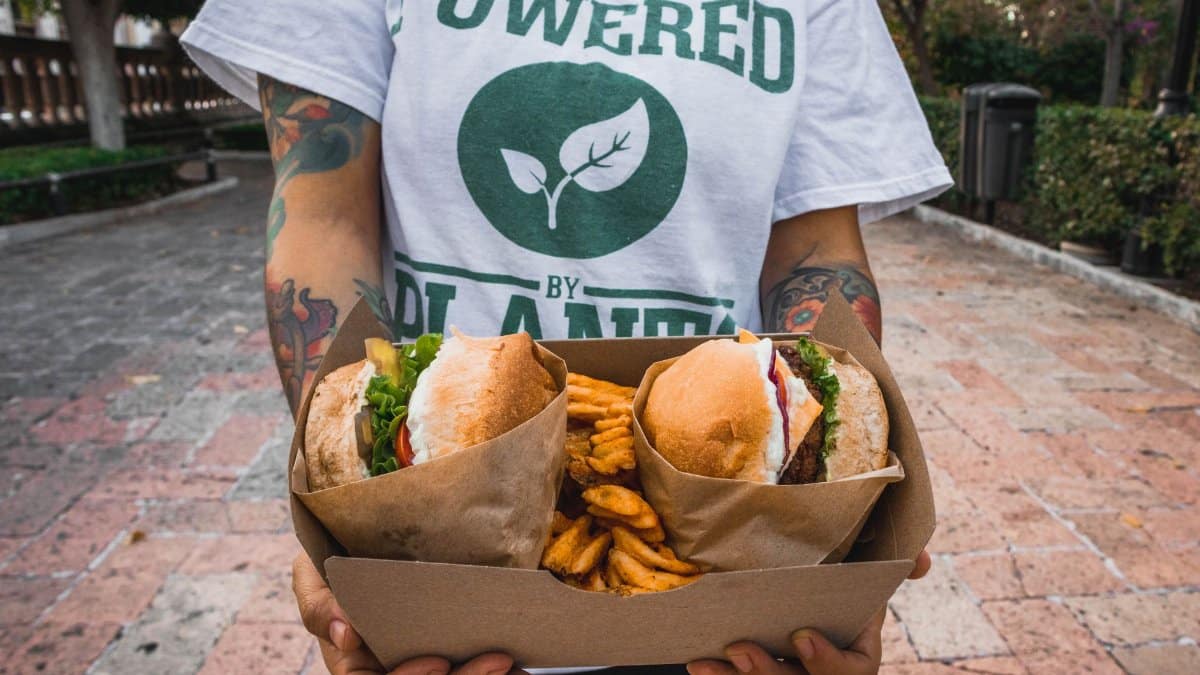 Person holding a takeout box with vegan burgers and fries, featuring tattoos and a plant-based message shirt, outdoors in Aguascalientes.