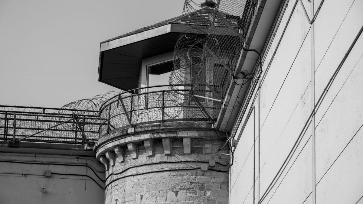 Black and white image of a prison tower secured with barbed wire fencing.