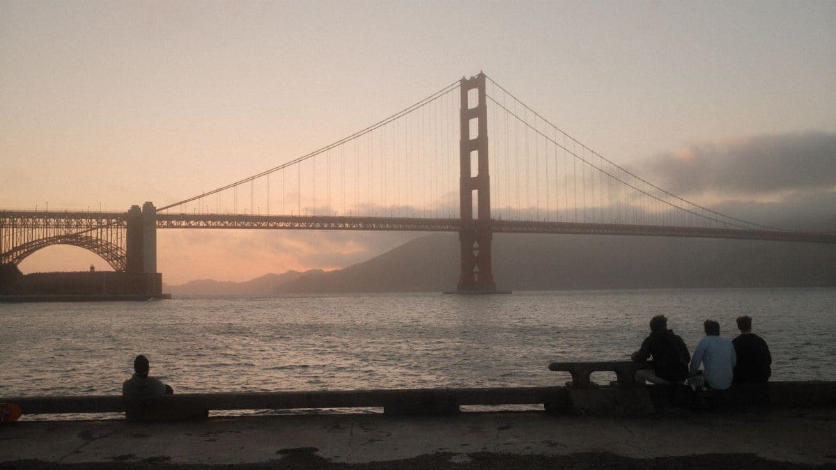 Golden Gate Bridge silhouetted at sunset, viewed from San Francisco with four people admiring the scene.
