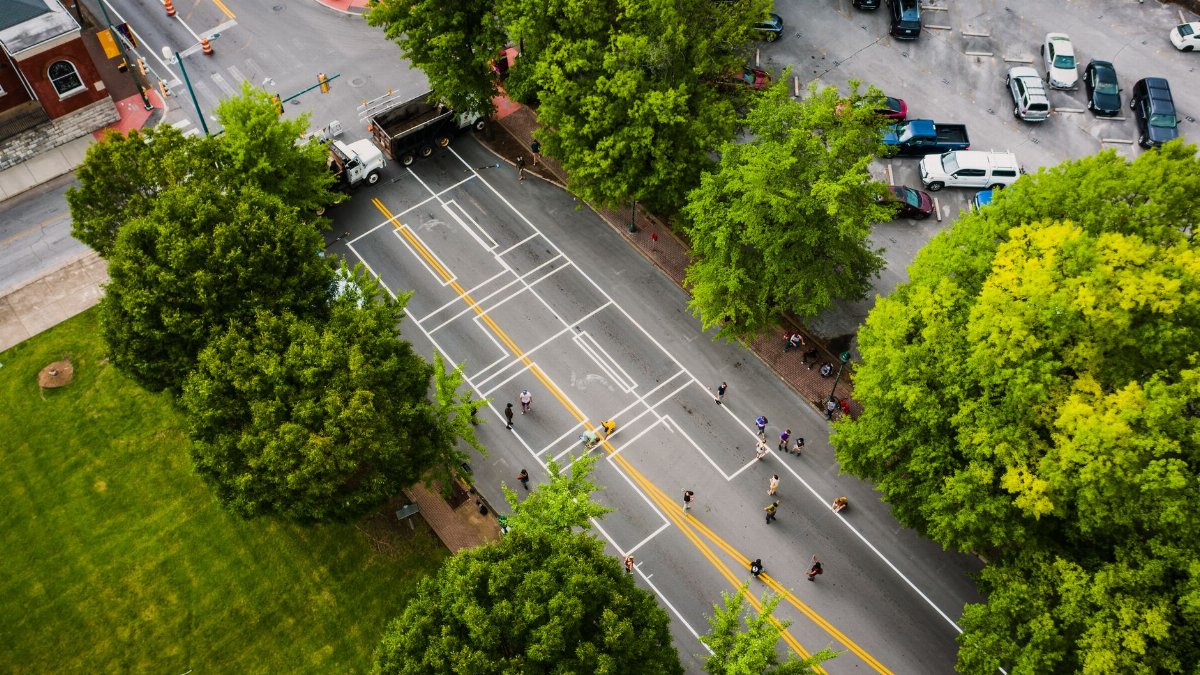 Aerial view of roadway with marking lines and unrecognizable people between meadow with bright trees and parking in city