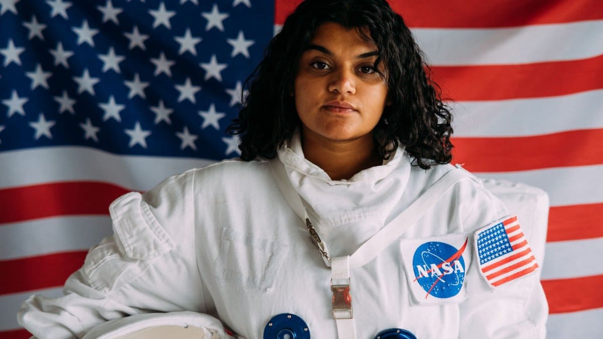 Woman in a NASA space suit standing in front of an American flag, representing exploration.