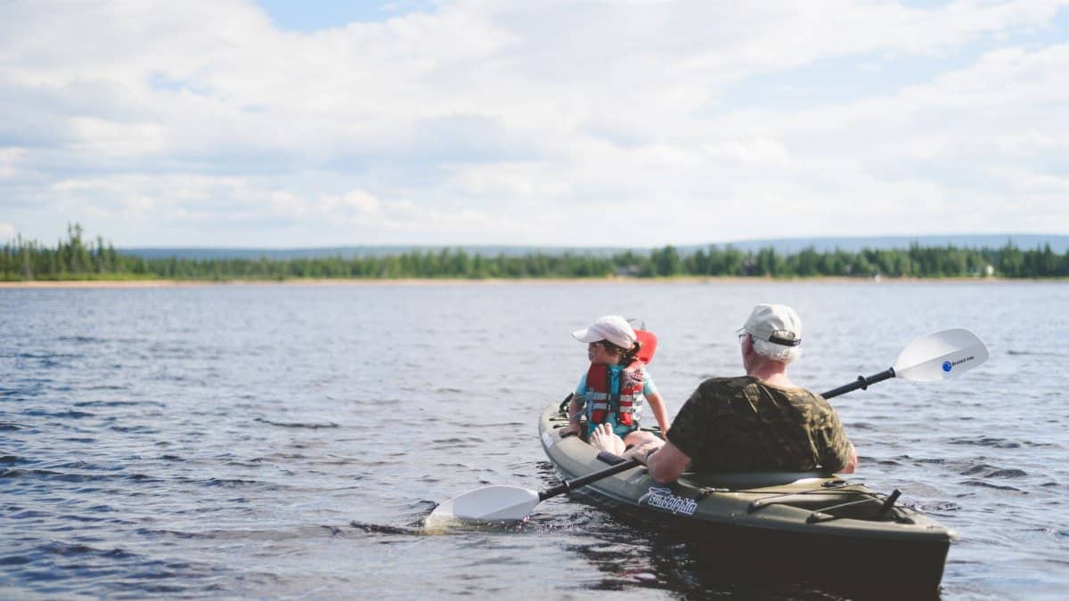 A grandfather and grandson kayaking together on a serene lake under a blue sky, enjoying outdoor adventure.