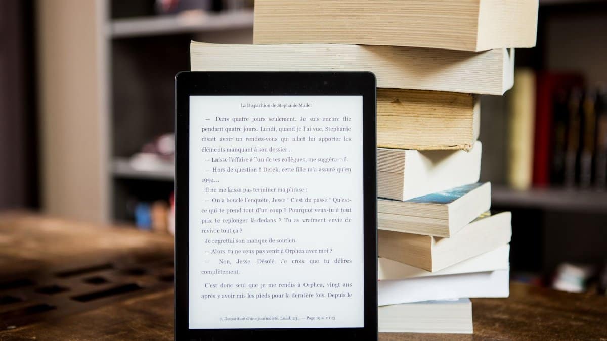 E-reader with text displayed next to a stack of physical books on a wooden table.