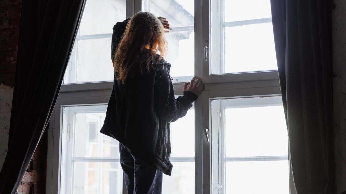 A woman stands by a window, lost in thought, with natural light streaming in.